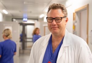 Läkaren Lars Ny i en korridor där andra vårdklädda personer hastar förbi i bakgrunden