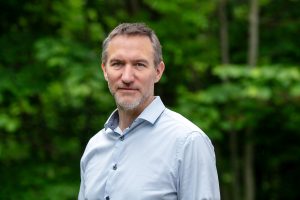 Man med skägg och ljus skjorta står framför skog.